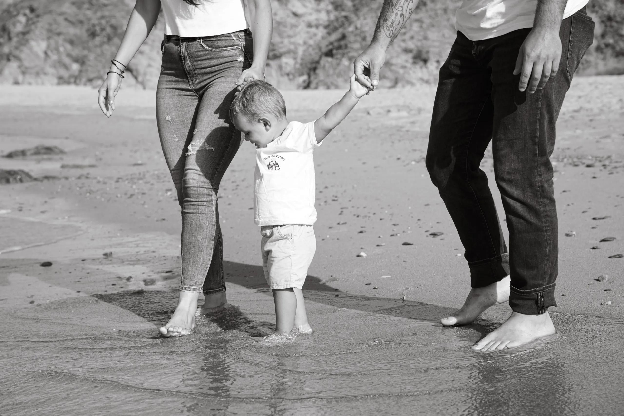 Un enfant fait ses premiers pas dans l’eau, main dans la main avec ses parents, sur une plage de Bretagne Sud